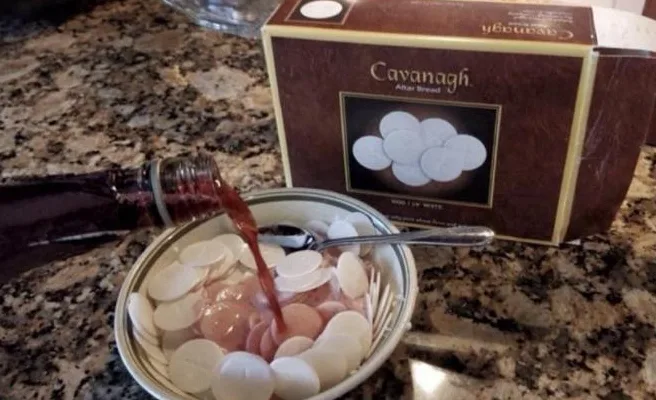 A bowl filled with white, round, flat discs labeled 'Cavanagh Alka Bread' sits on a granite countertop. A dark red liquid is…
