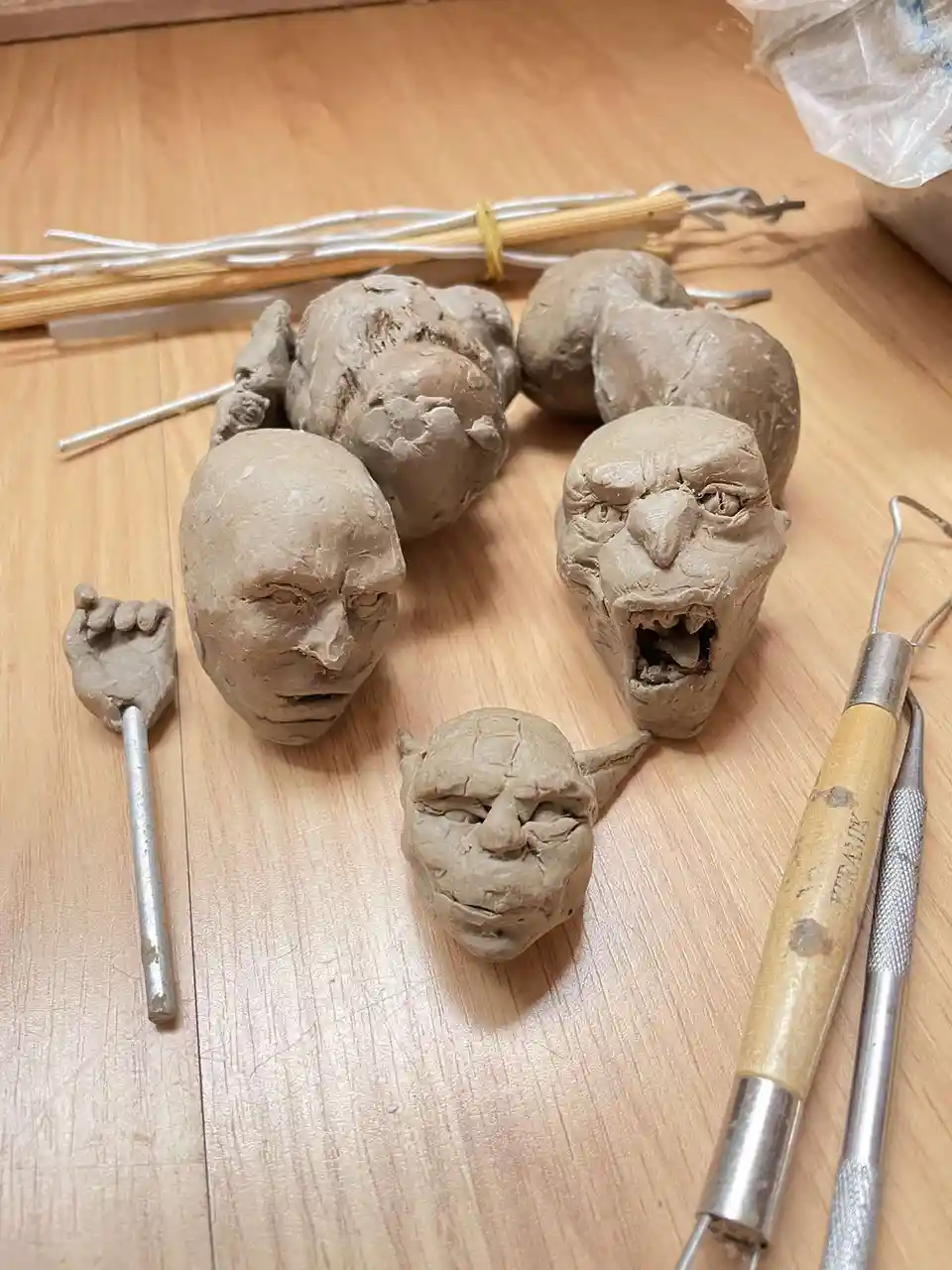 Several unfinished clay sculptures of human and demonic faces sit on a wooden table, surrounded by sculpting tools like loop…
