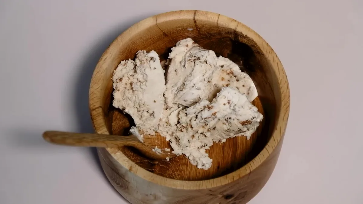 A wooden bowl filled with scoops of vanilla ice cream with chocolate chunks, with a wooden spoon resting inside. The image…