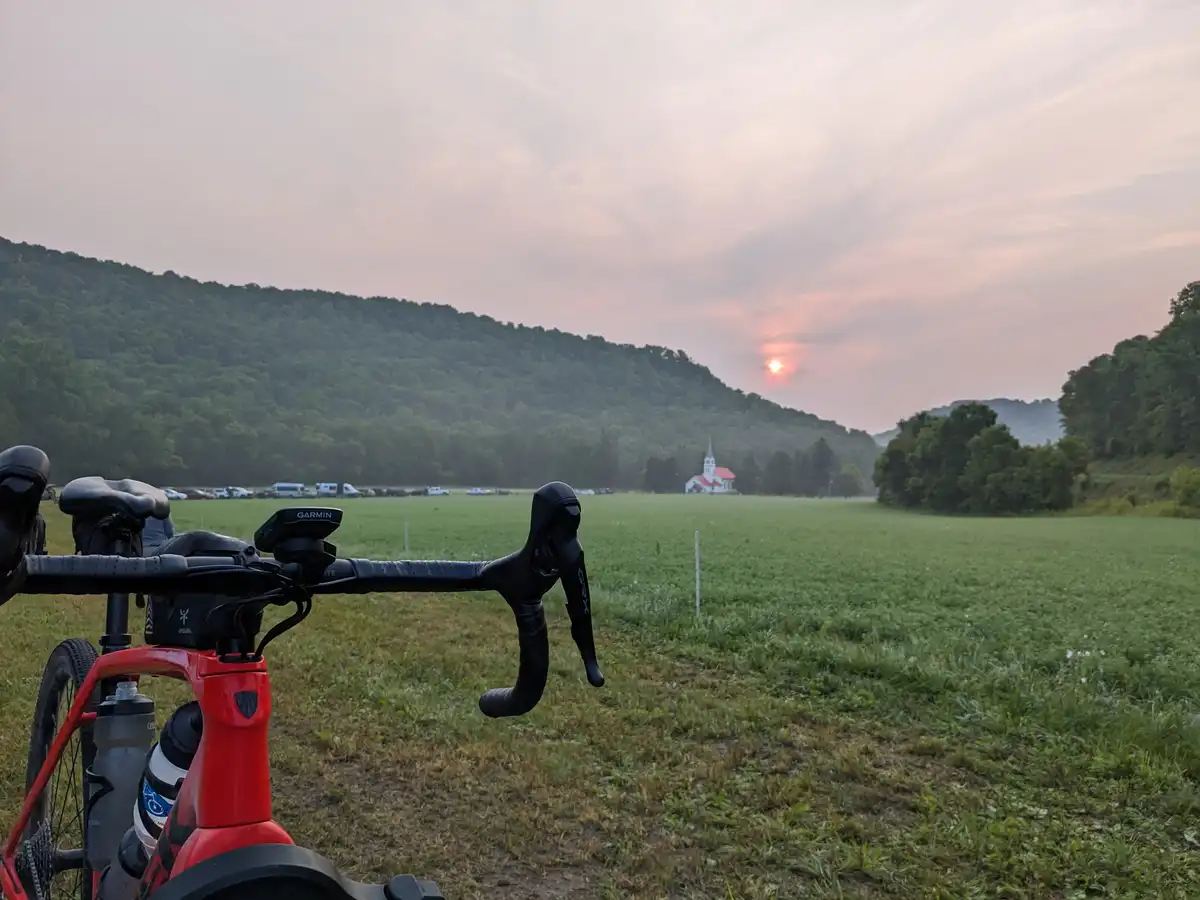 A first-person view from a red road bike with black handlebars and a Garmin computer, parked in a grassy field at sunset. In…