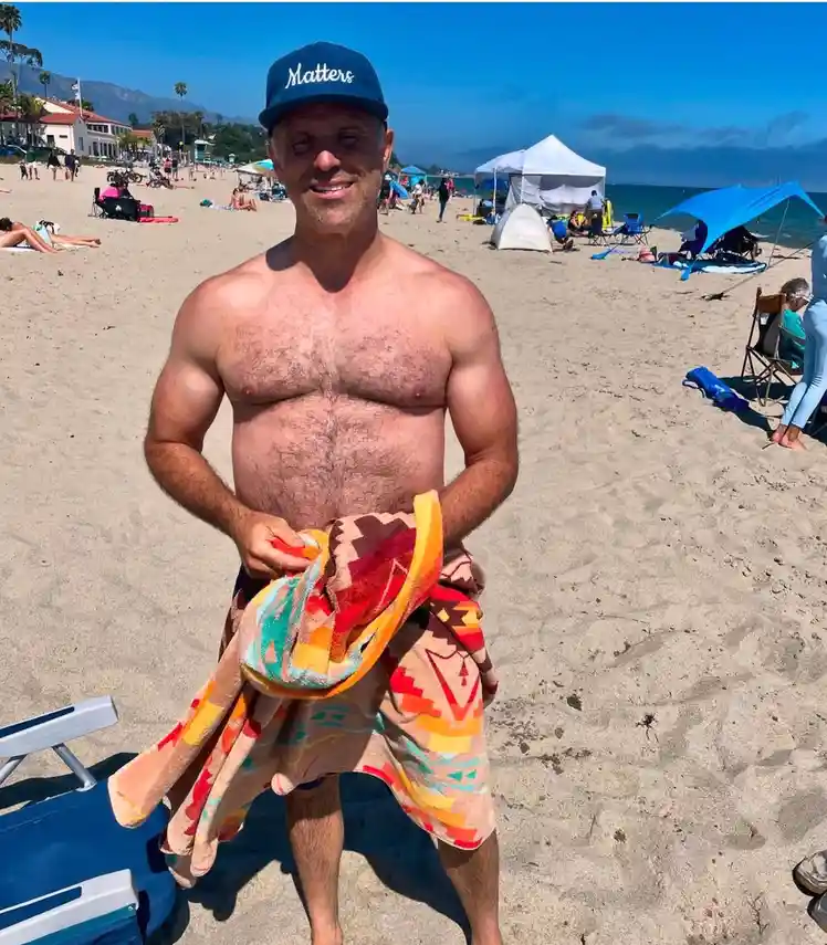 A shirtless man with a hairy chest stands on a sunny beach, wearing a blue cap with the word 'Matters' written on it and…
