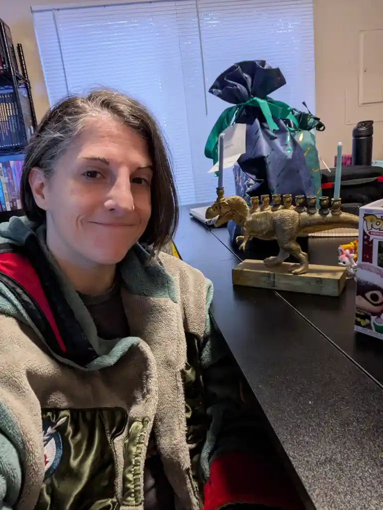 A woman with short dark hair and gray streaks smiles at the camera while wearing a fleece jacket. Behind her on a black…