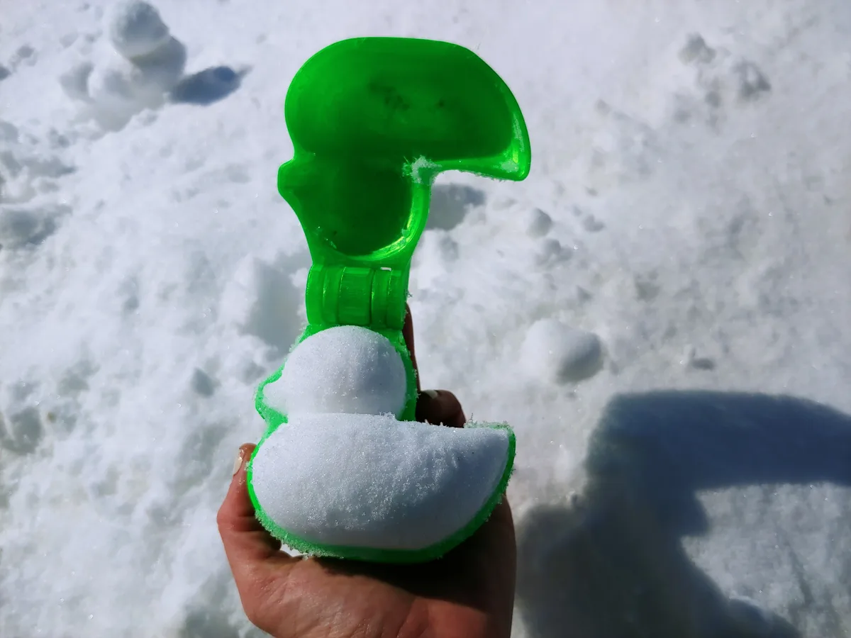A hand holds a green plastic snow mold shaped like a question mark, filled with packed snow, against a snowy background. The…
