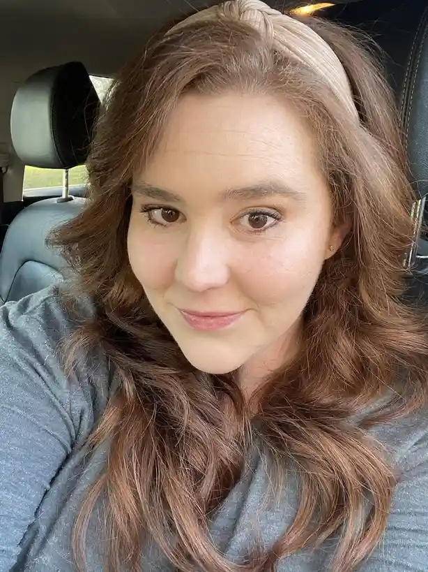 A woman with long brown hair and a beige headband sits in a car, looking directly at the camera with a tired but slightly…