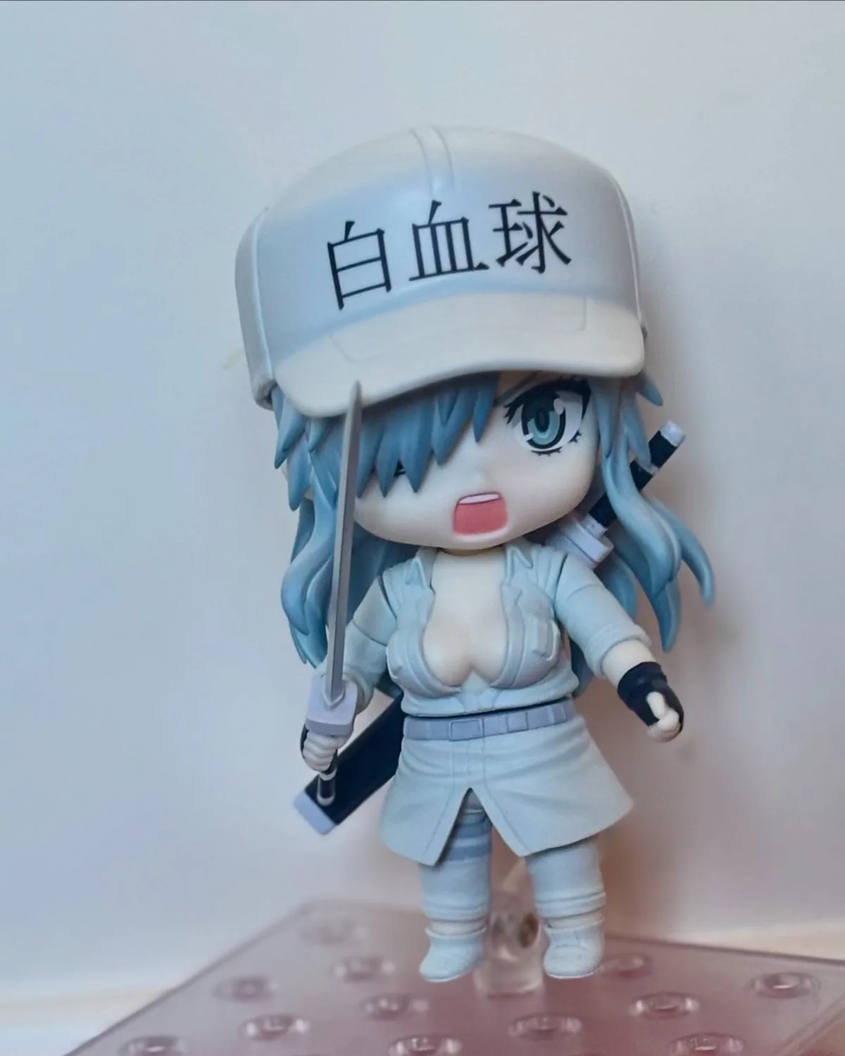 A chibi-style anime figurine of a female character with blue hair and large eyes, wearing a white cap with the Japanese text…