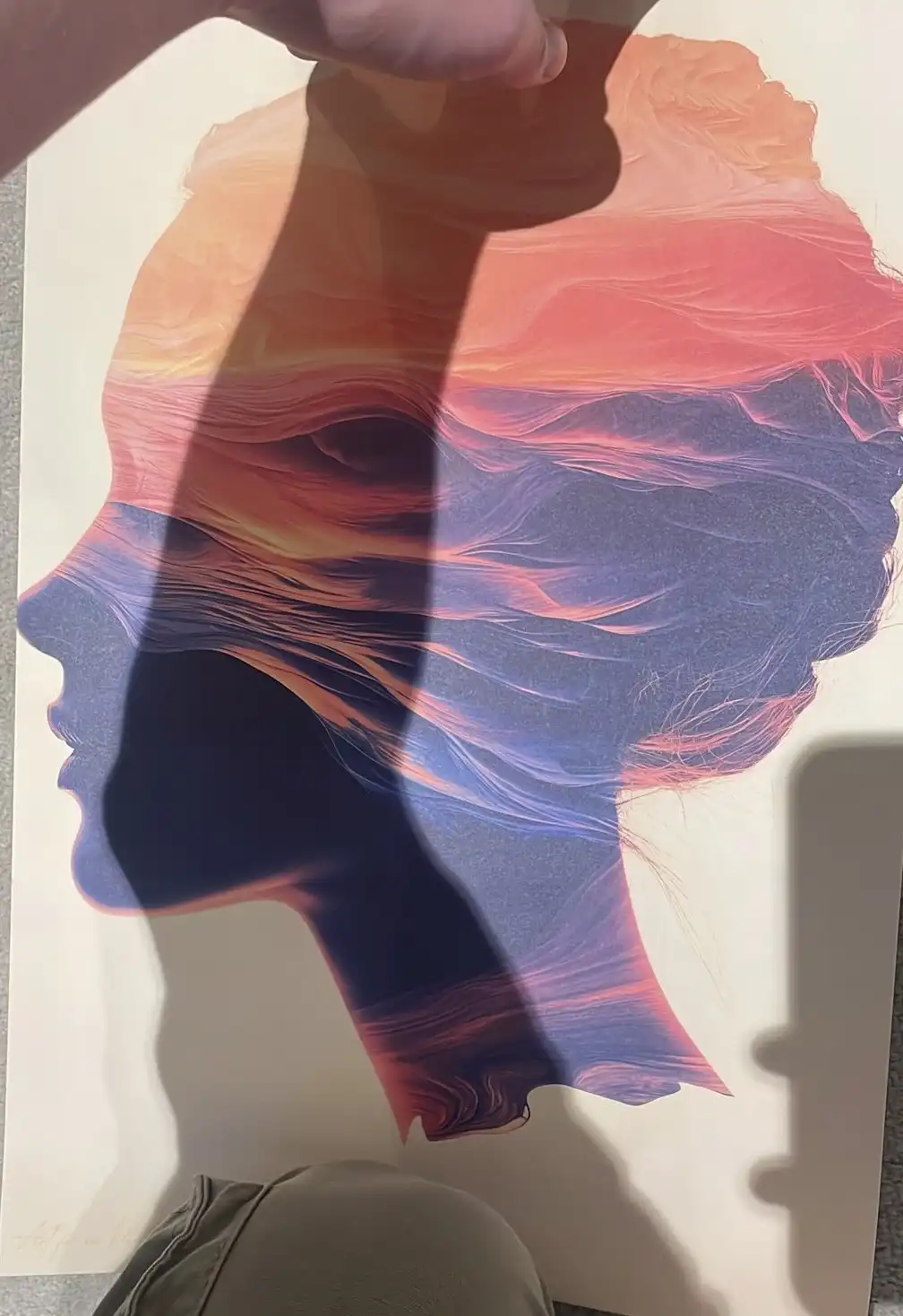 A person holds up a colorful art print depicting a stylized human profile in silhouette, filled with swirling gradients of…