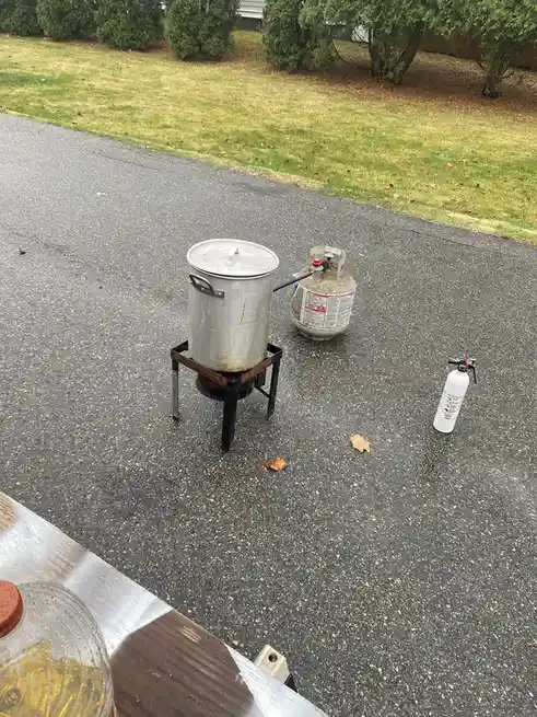 A large metal pot sits on a portable burner on an asphalt driveway, connected to a propane tank. A white fire extinguisher…