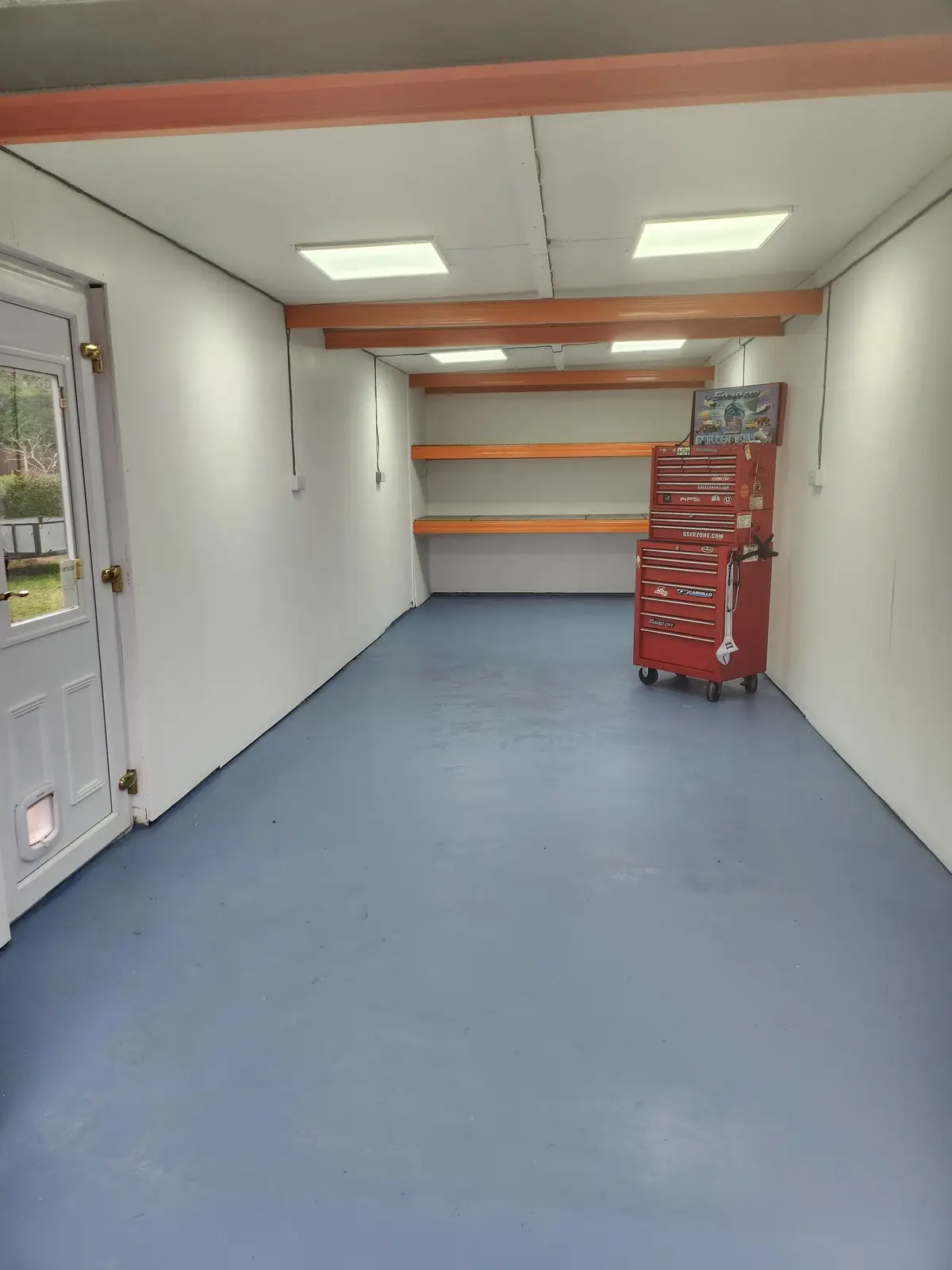 An empty, clean garage with white walls, a blue floor, and orange trim on beams and shelves. A single red rolling tool chest…