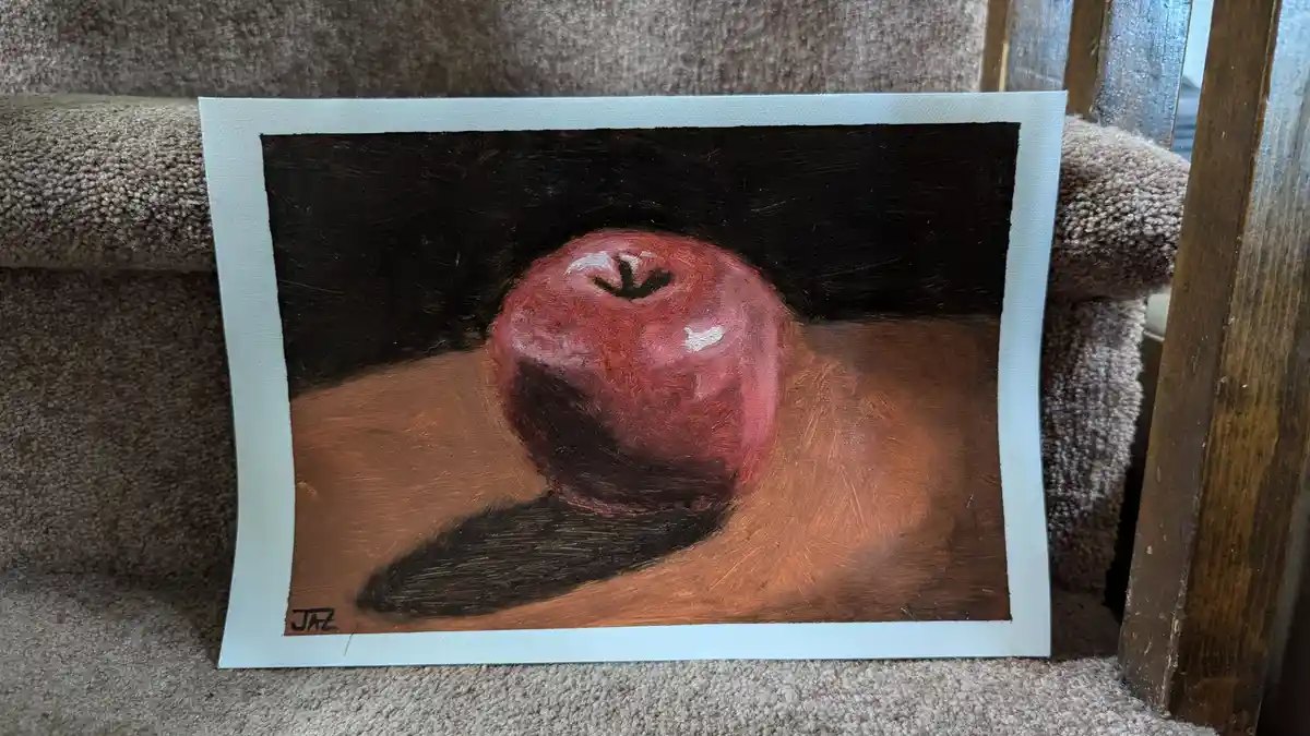 A painting of a red apple on a brown surface against a black background, signed 'JAZ' in the bottom left corner, is propped…