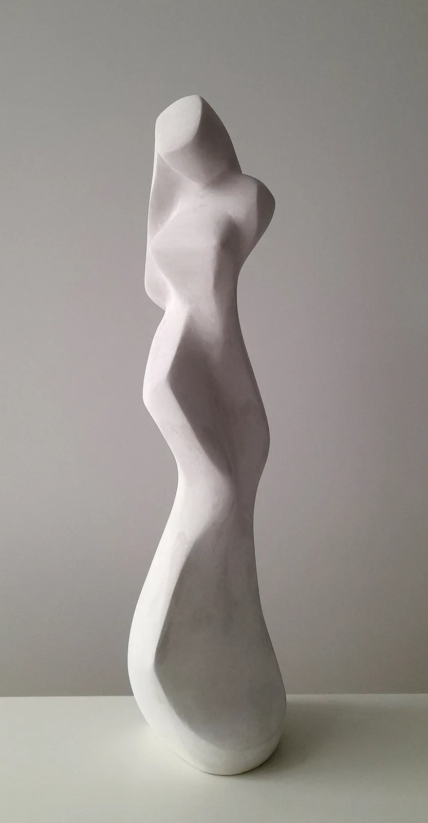 A minimalist white abstract sculpture of a human torso and legs, viewed from the side against a plain gray background. The…