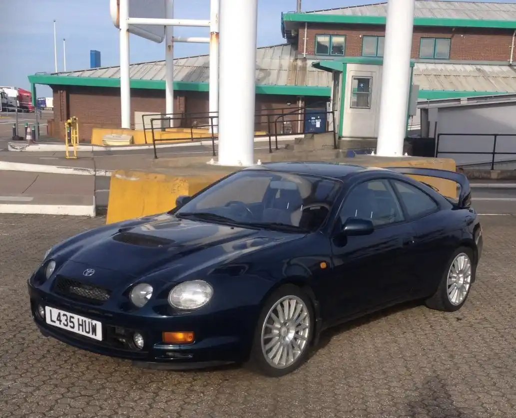 A dark blue Toyota Celica GT-4 is parked at a toll booth or service station, with its license plate clearly visible as 'L435…