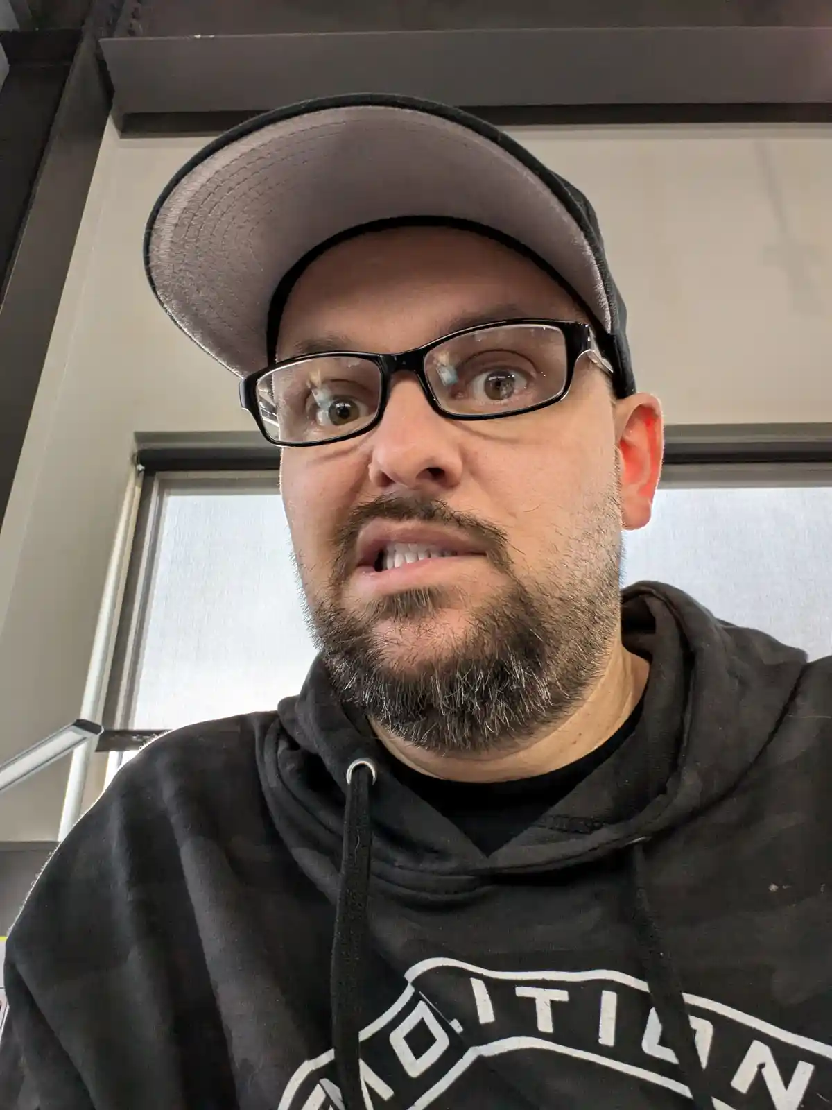 A man with glasses and a backward cap stares with wide, shocked eyes and an open mouth, wearing a black hoodie with white…