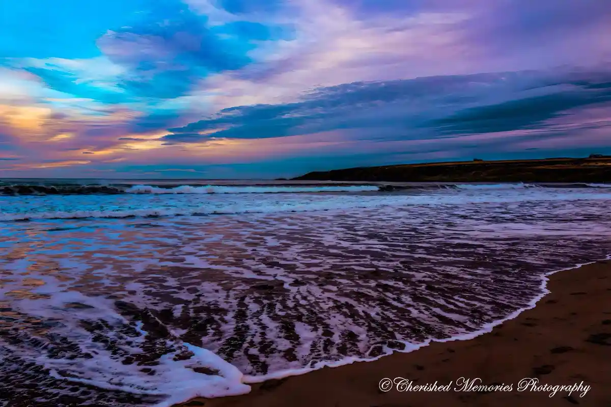 A serene beach scene at sunset or sunrise with vibrant blue, purple, and orange clouds reflected in the ocean waves rolling…