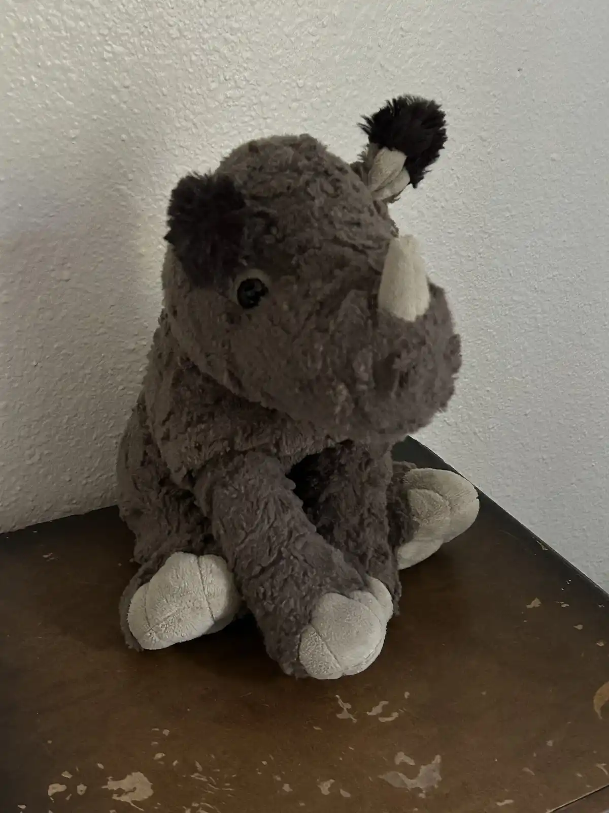 A gray plush rhino toy with black ears and white hooves sits on a worn wooden surface against a textured white wall. The…