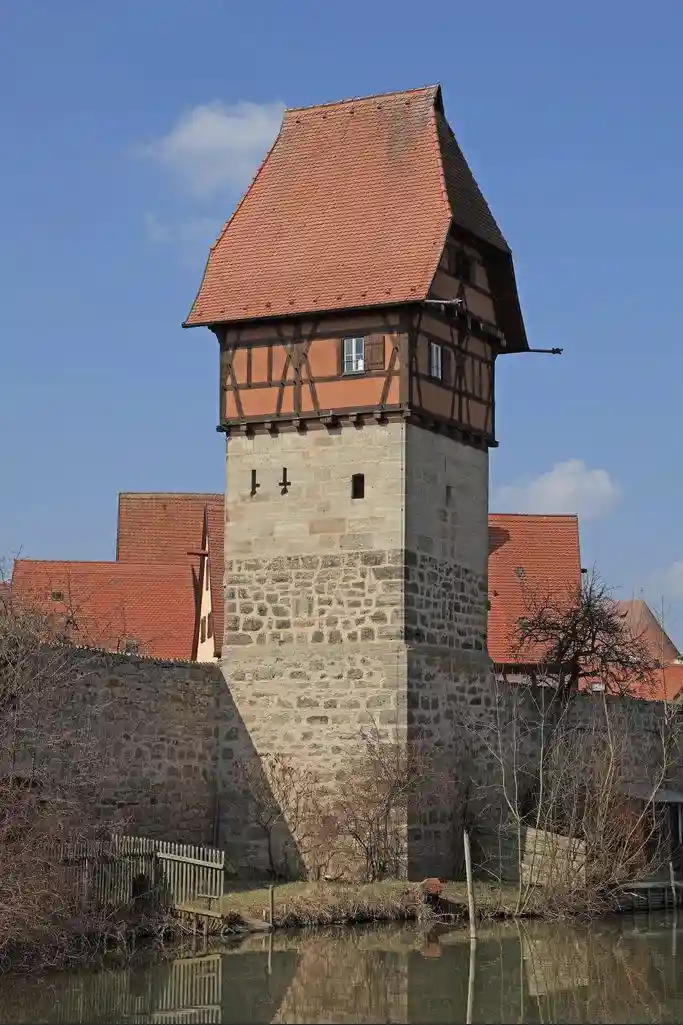 A tall, historic stone and timber-framed tower with a red-tiled roof stands beside a moat under a blue sky. The image has…