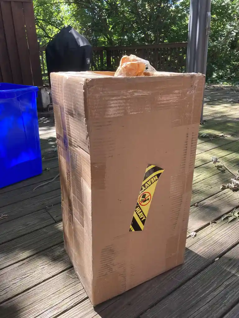 A large, tall cardboard shipping box sits on a wooden deck, featuring a prominent yellow and black 'HEAVY' warning sticker…