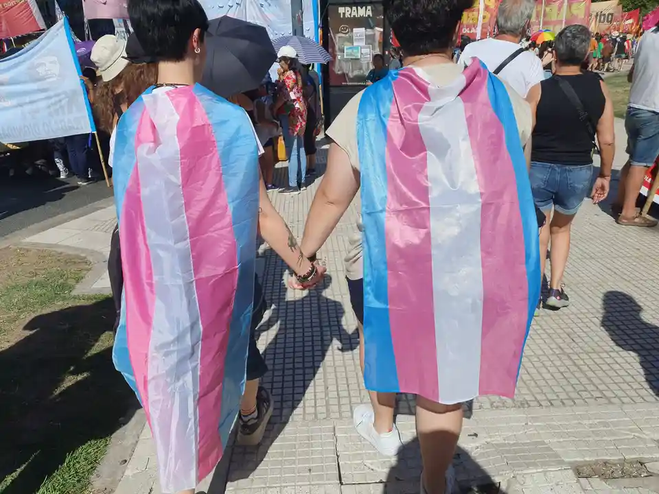 Two people are seen from behind walking hand-in-hand down a paved sidewalk at an outdoor gathering, both draped in trans…