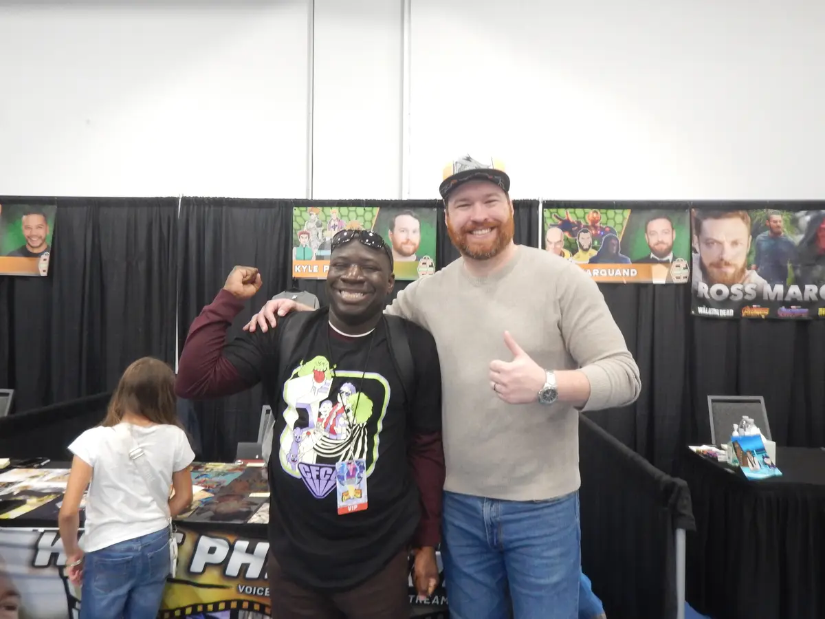 Two men pose for a photo at a convention booth with posters of voice actors behind them. The man on the left, wearing a…