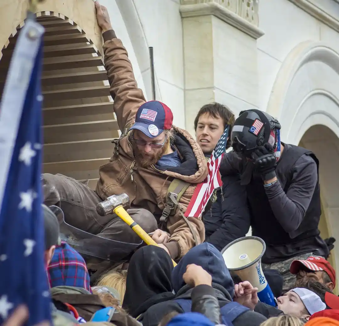 A man with a beard and glasses, wearing a red, white, and blue 'Trump 2020' cap, sits on steps while holding a…