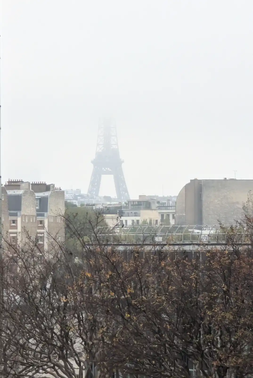 A hazy, overcast photograph shows the Eiffel Tower barely visible through thick fog, with urban buildings and bare-branched…