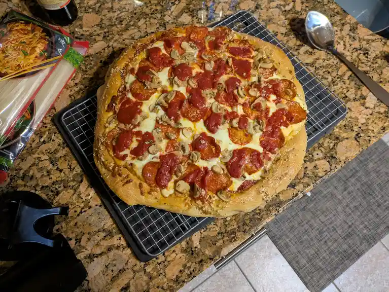 A homemade pepperoni and mushroom pizza sits on a cooling rack on a granite countertop, surrounded by a package of Asian…