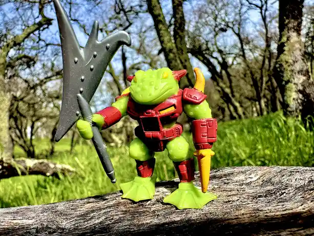 A green plastic toy figure resembling a frog with red armor and horns stands on a log in a grassy outdoor setting, holding a…