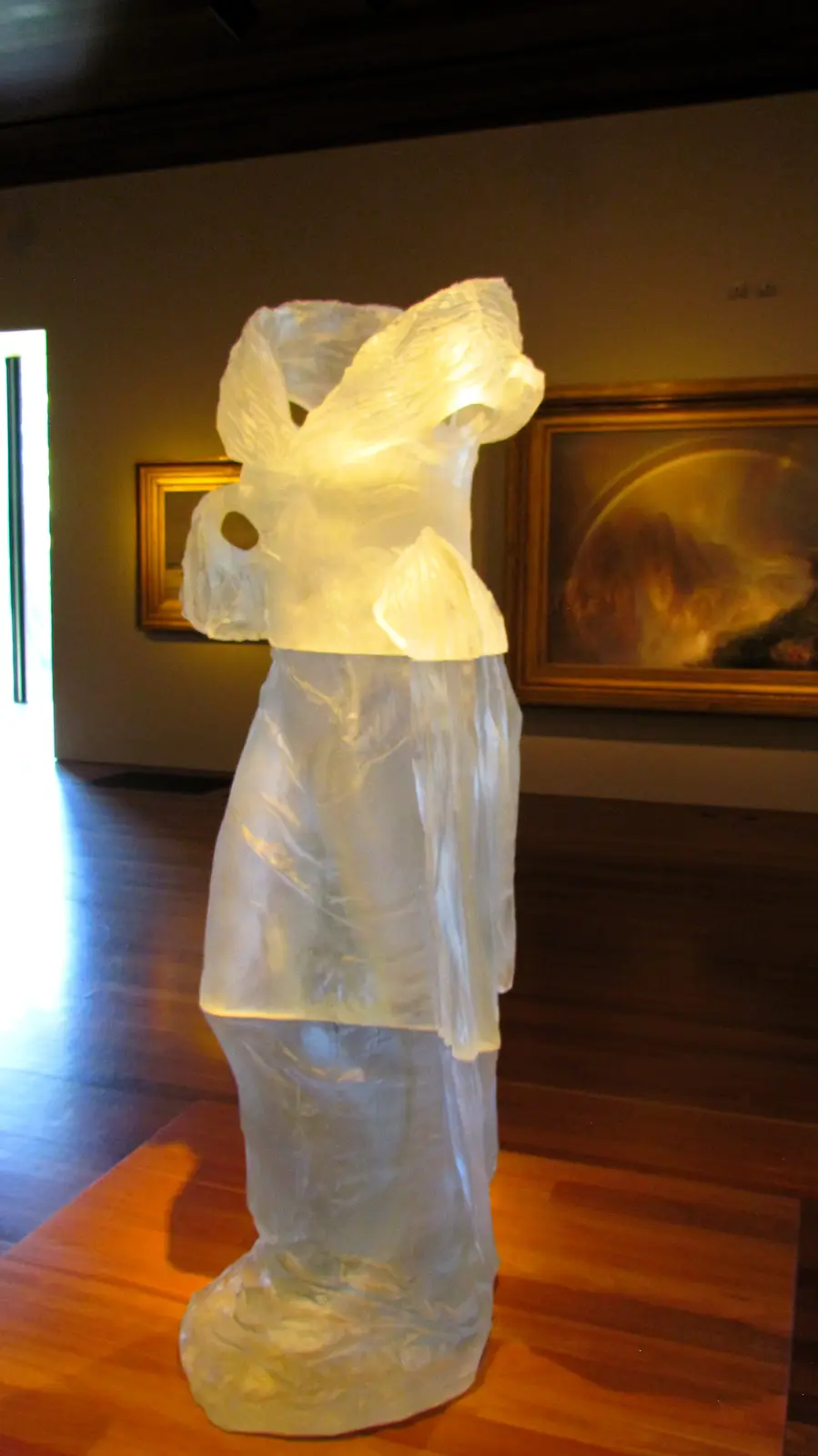 A translucent, glowing sculpture shaped like a human figure stands on a wooden pedestal in an art gallery, with framed…