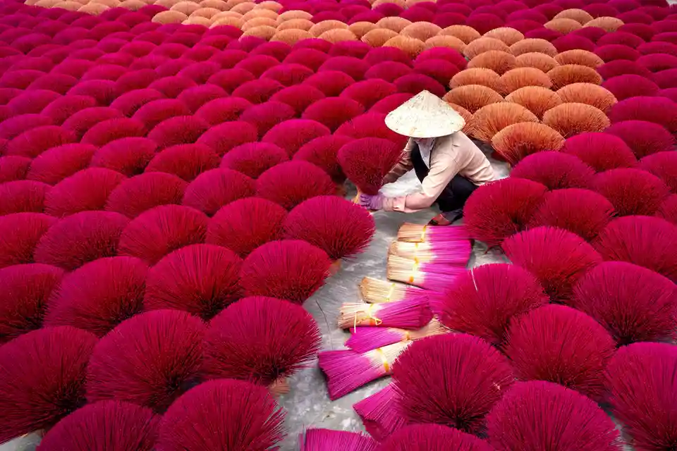 A person wearing a traditional conical hat is crouched among vast rows of brightly colored incense sticks arranged in fan…