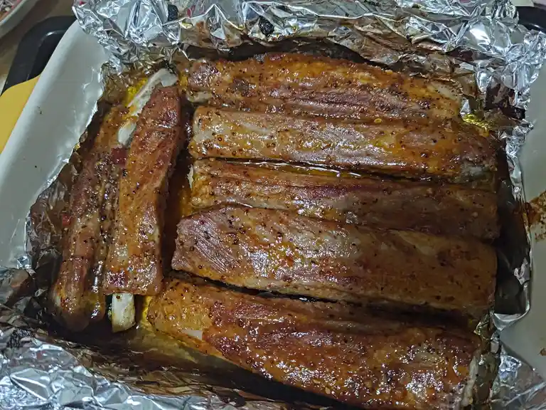 A close-up photo shows several cooked ribs resting on aluminum foil inside a white baking dish. The ribs are glazed with a…