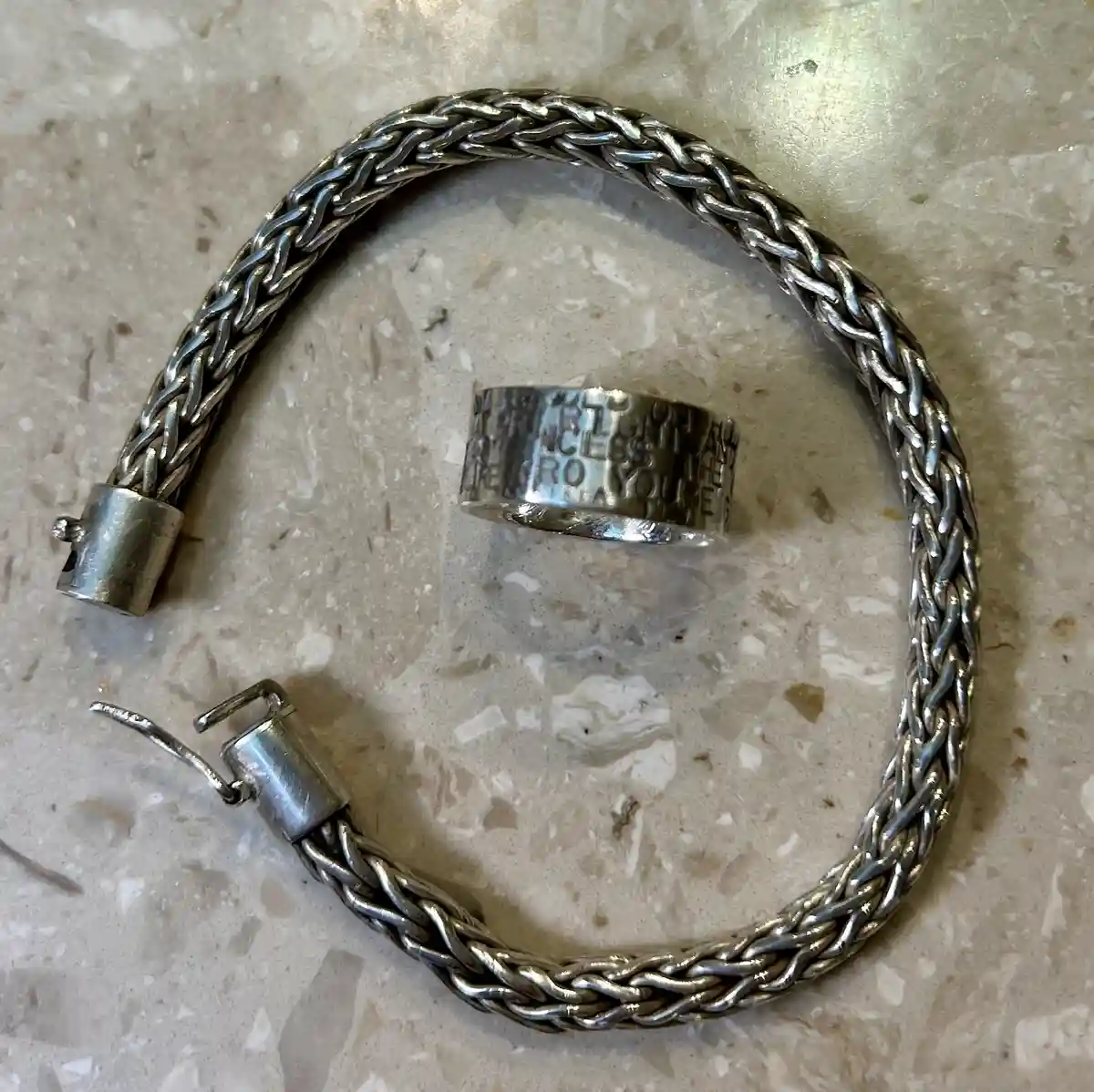 A silver braided bracelet with a magnetic clasp lies on a speckled beige countertop, with a wide silver ring inscribed with…