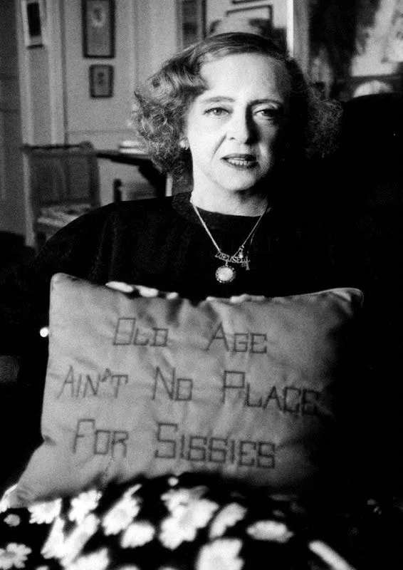 A black-and-white photograph of a woman with curly hair holding a pillow that reads 'Old Age Ain't No Place For Sissies'…