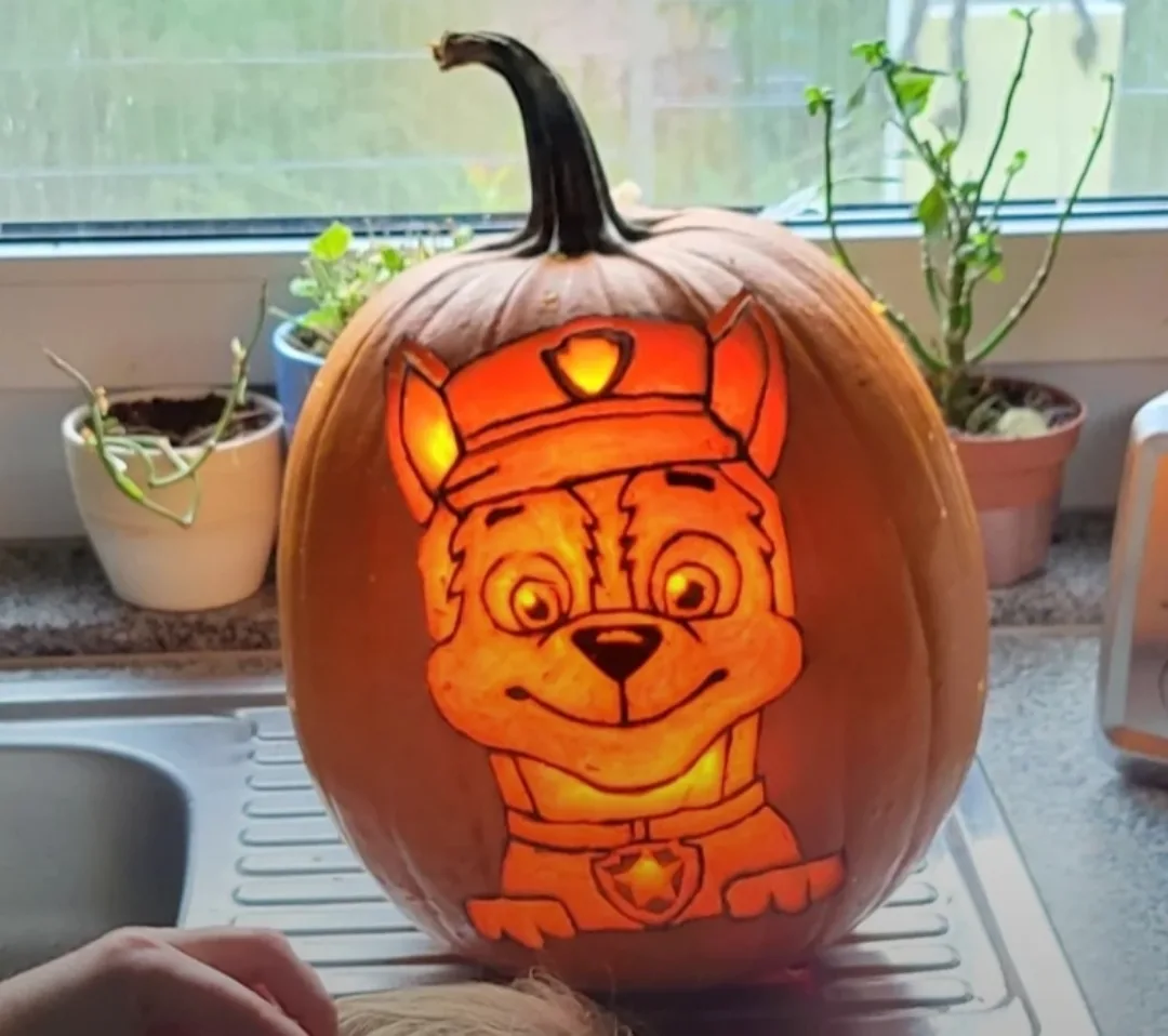 A pumpkin is intricately carved with the face of Chase, the police dog from Paw Patrol, and is illuminated from within…