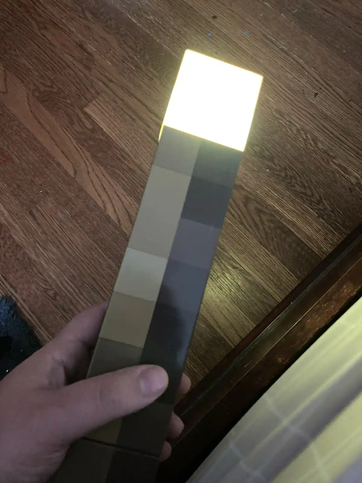 A hand holds a Minecraft-style torch with a bright, glowing top and a pixelated, blocky body made of gray and beige squares…