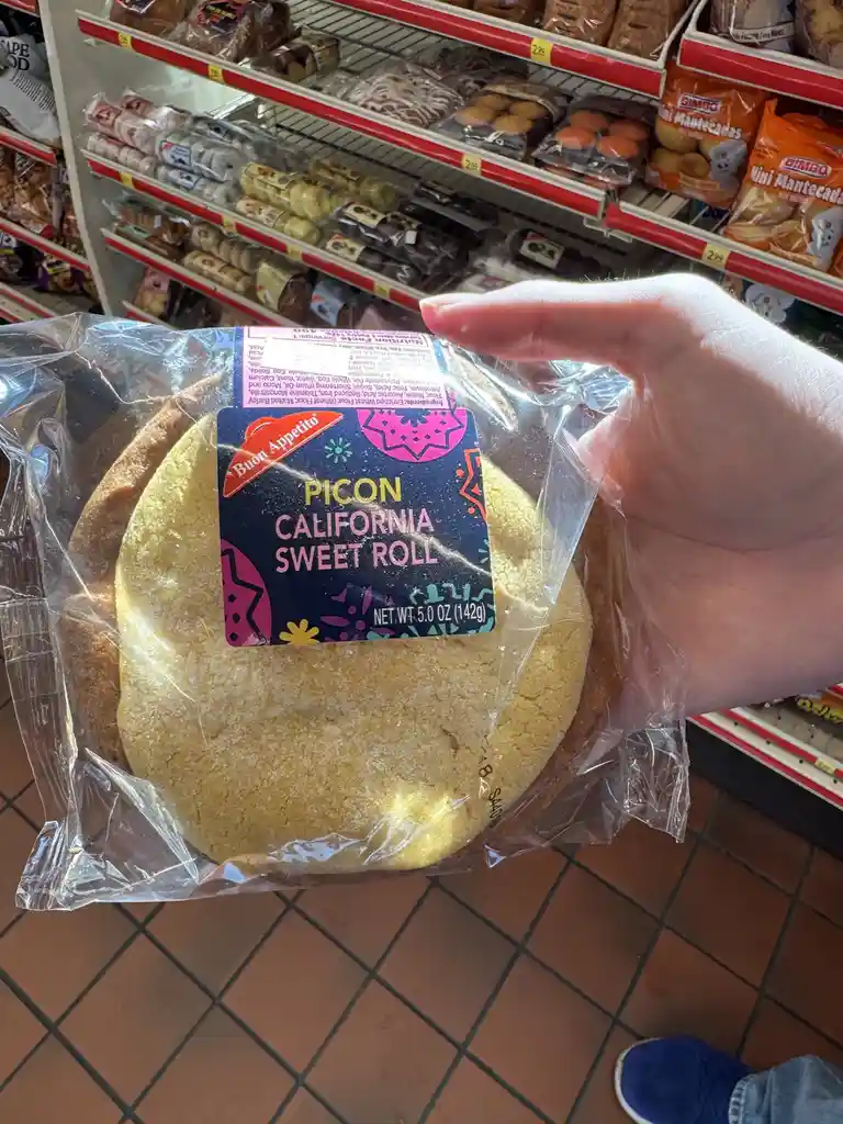 A hand holds a large, round, yellow cake-like pastry in clear plastic packaging labeled 'PICON CALIFORNIA SWEET ROLL' in a…