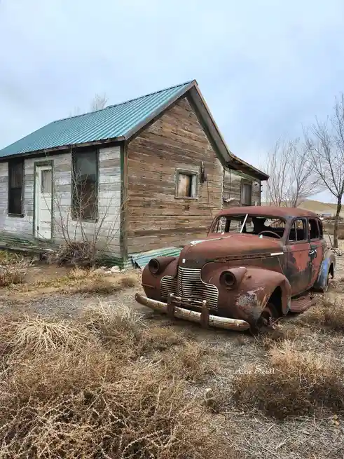 A rusted, abandoned vintage car sits in front of a dilapidated wooden house with a green metal roof, surrounded by dry brush…