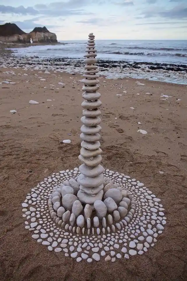 A tall, meticulously stacked tower of smooth, rounded stones stands on a sandy beach, surrounded by concentric circles of…