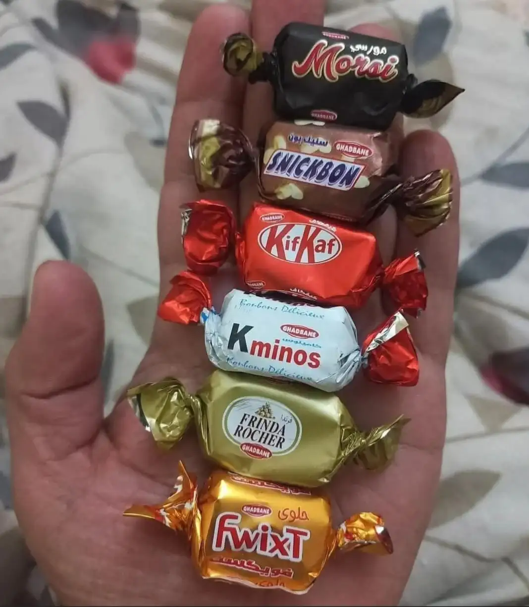 A hand holds a vertical stack of six small, individually wrapped candies from Ghadbane, including Morai, SnickBon, KifKaf…