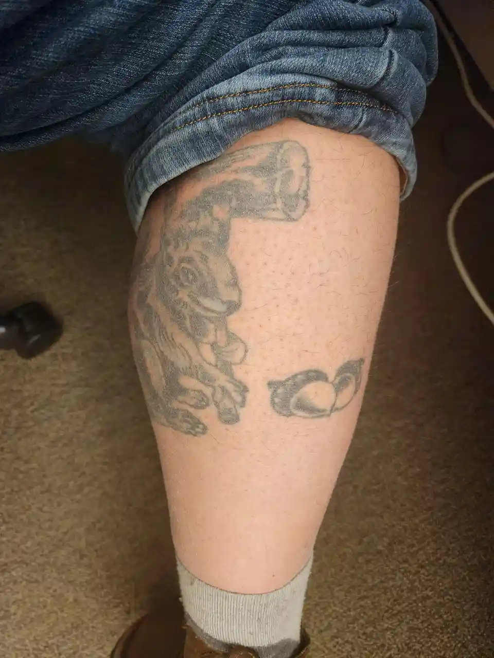 A close-up photo shows a person's lower leg with rolled-up blue jeans, revealing a faded black-and-gray tattoo of a hammer…
