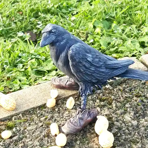A statue of a black raven stands on a gravel path next to a low stone border with green grass behind it. The raven is…