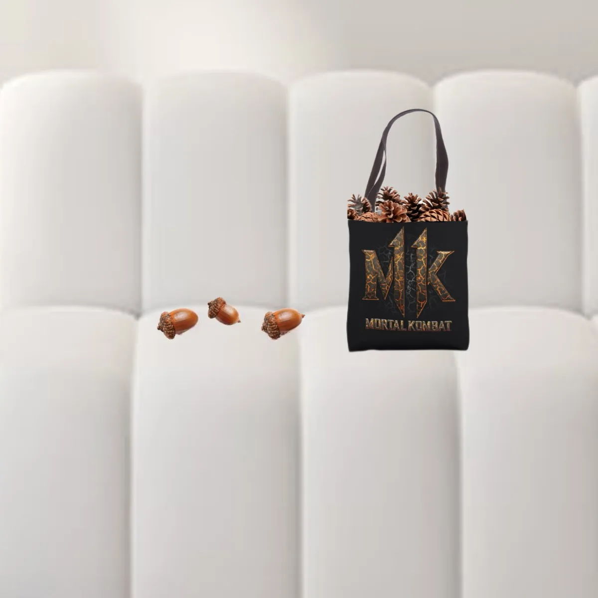 A black 'Mortal Kombat' tote bag filled with pinecones sits on a white tufted couch, with three acorns scattered nearby on…