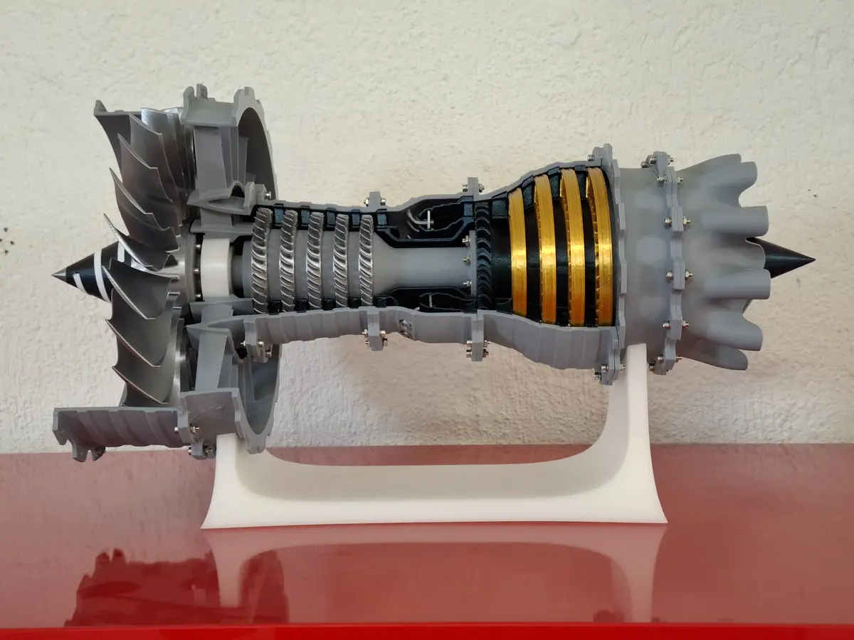 A 3d printed cross-section model of a jet engine is displayed on a white stand against a textured wall, revealing its…