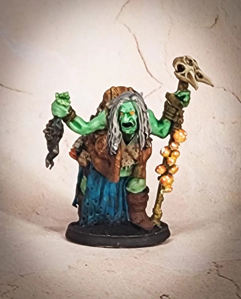 A painted miniature figurine of a green-skinned, long-haired witch with an angry expression, wearing a brown vest and blue…