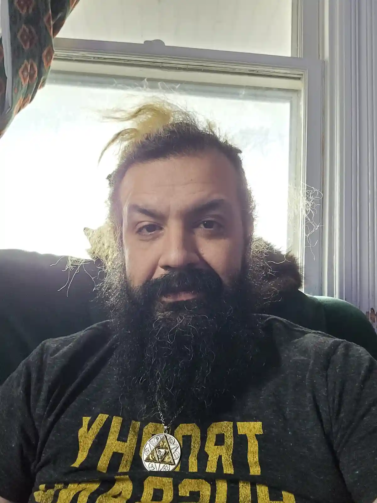 A man with a long dark beard and messy hair sits in front of a window, wearing a black t-shirt with yellow text that appears…