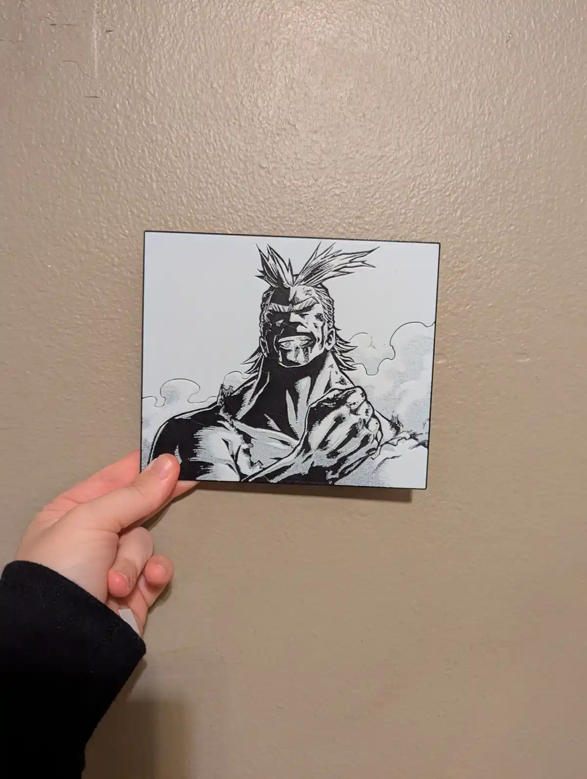 A hand holds up a square, black-and-white printed image of the anime character All Might from My Hero Academia, who is…