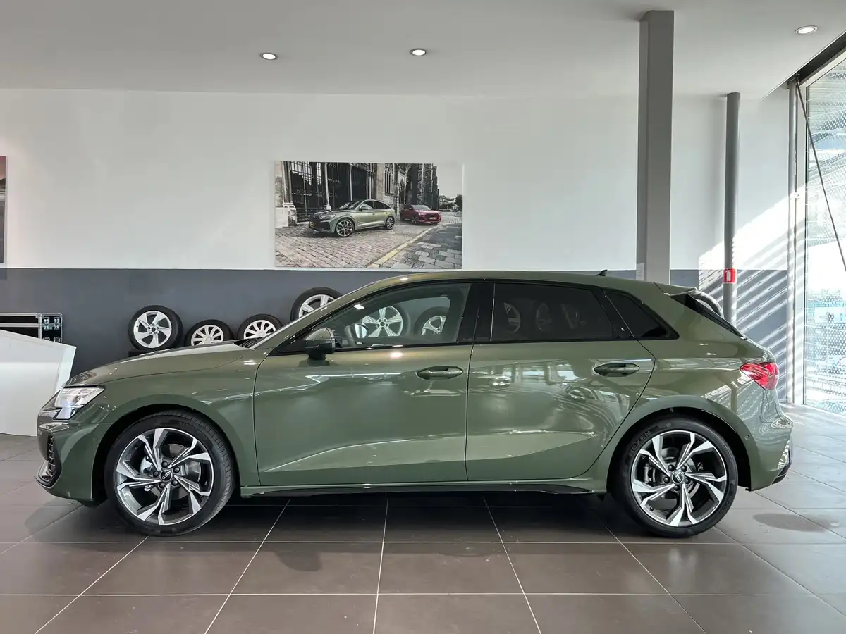 A green Audi A3 Sportback is parked inside a modern car dealership showroom, with a large promotional poster of the same…