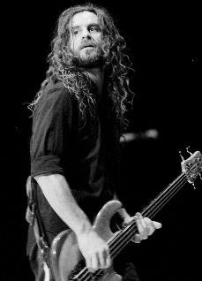 A black-and-white photo of a man with long curly hair and a beard, wearing a dark shirt and playing a bass guitar on stage…