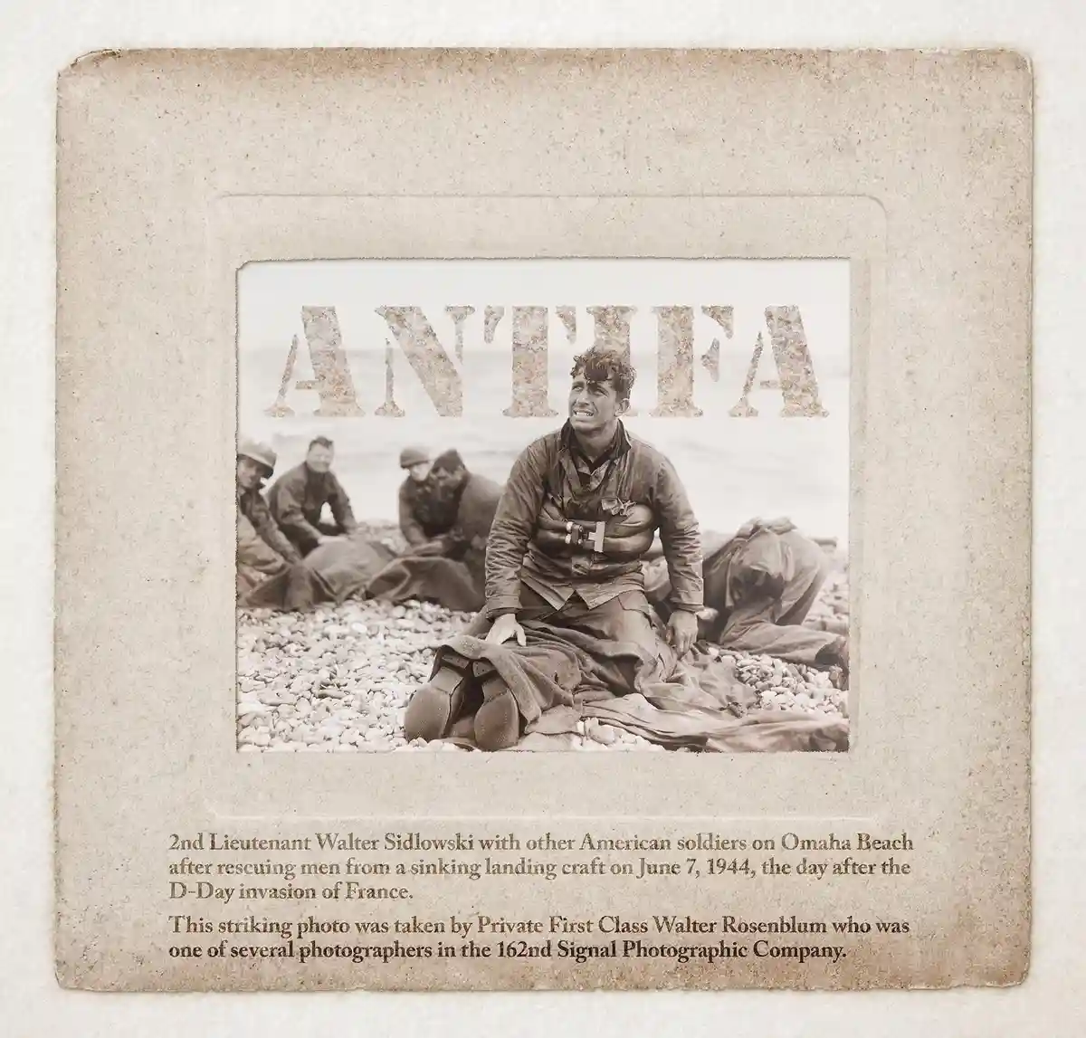 A vintage-style photo shows a group of American soldiers resting on a pebbled beach, with one soldier in the foreground…