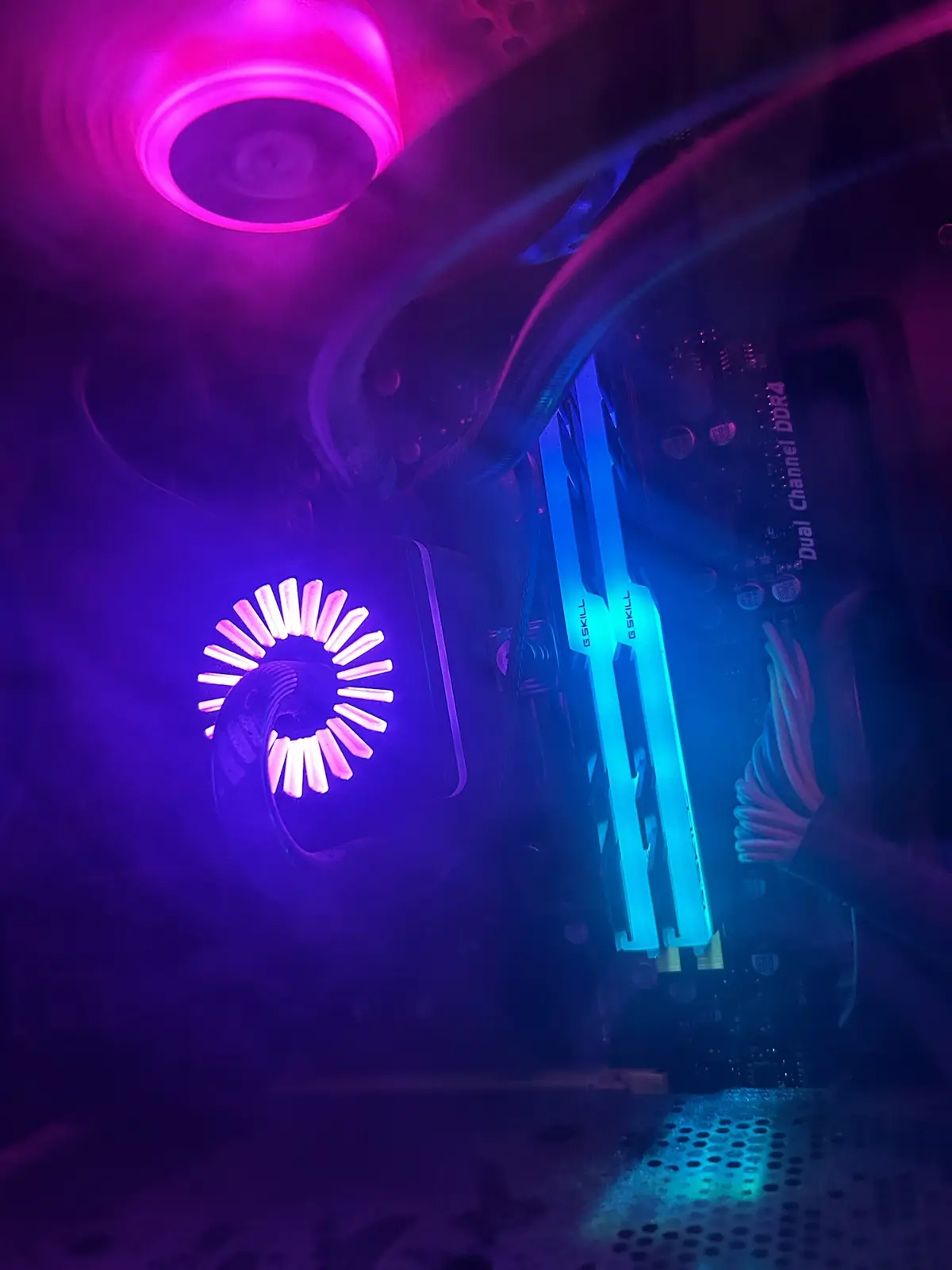 A close-up shot inside a computer case showing glowing pink and blue RGB lighting on a CPU cooler fan and G.SKILL RAM…