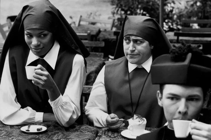 A black-and-white photo shows three people dressed as nuns sitting at a table with coffee cups and desserts. The nun on the…