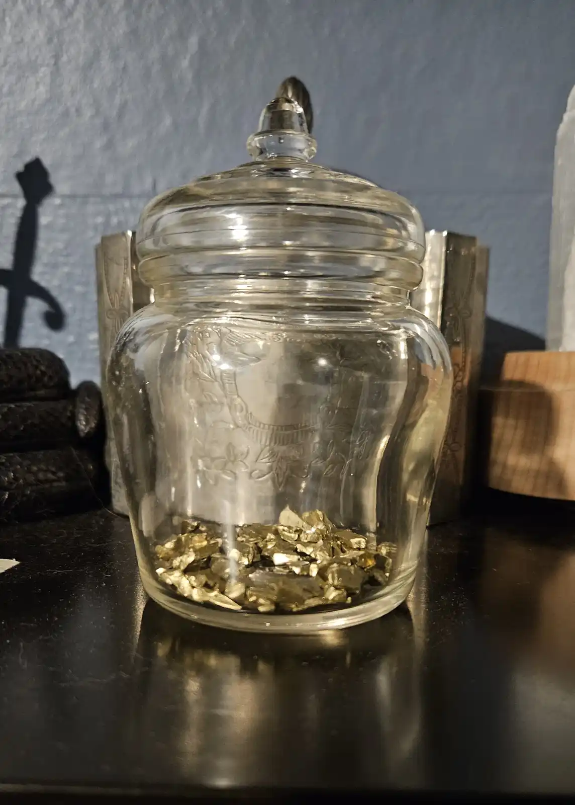 A clear glass jar with a lid, sitting on a dark surface, contains only a small amount of shiny, irregularly shaped…
